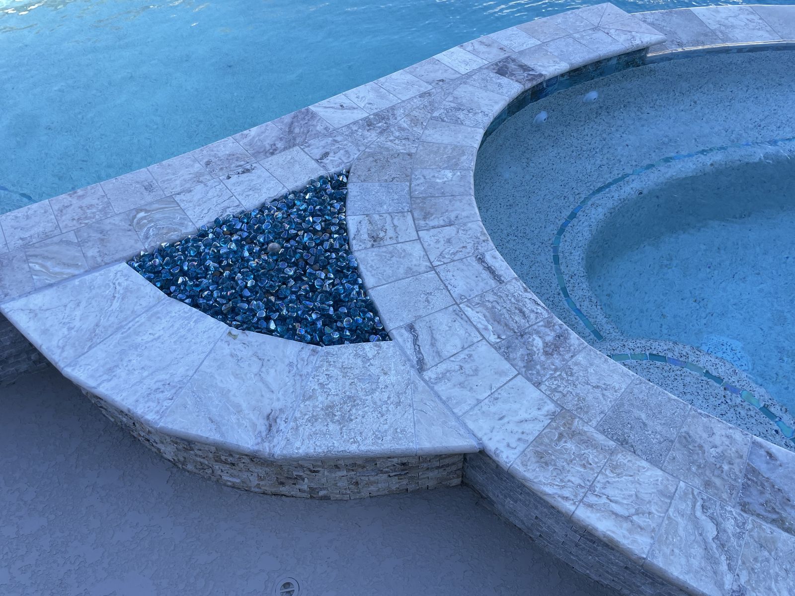 Fire bowl with blue glass set into travertine coping beside the spa