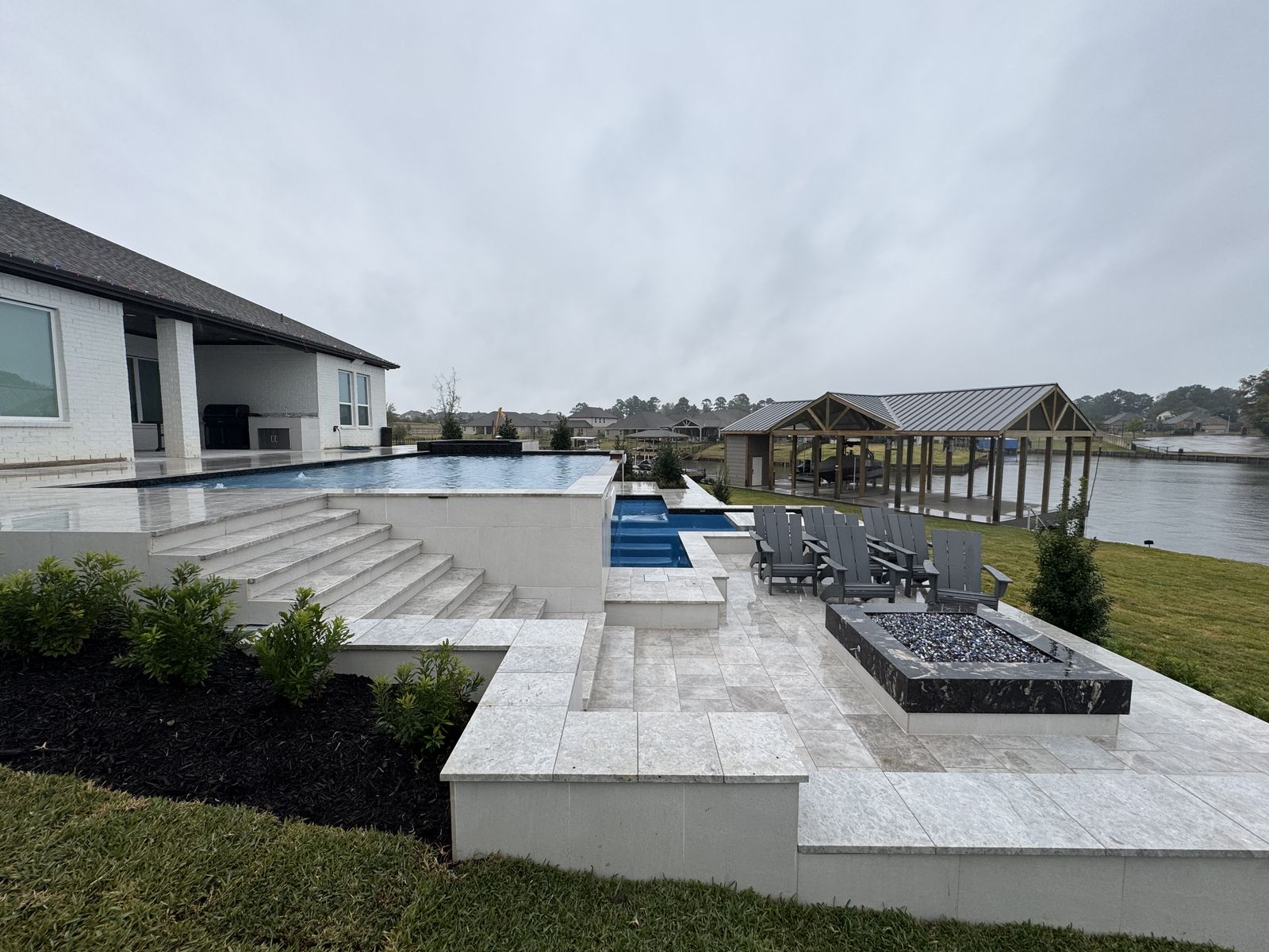 Infinity-edge lakefront pool with steps, fire pit and a boathouse behind it